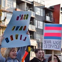 两个人举着标语:一个举着跨性别者的旗帜，上面写着“你是爱的”，另一个举着“GSA拯救生命”的标语。