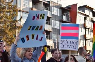 两个人举着标语:一个是写着“你被爱着”的跨性别标志，另一个是写着“GSA拯救生命”的标语。