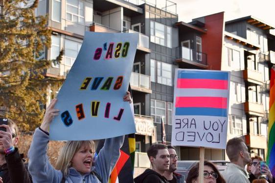 两个人举着标语:一个是写着“你被爱着”的跨性别标志，另一个是写着“GSA拯救生命”的标语。
